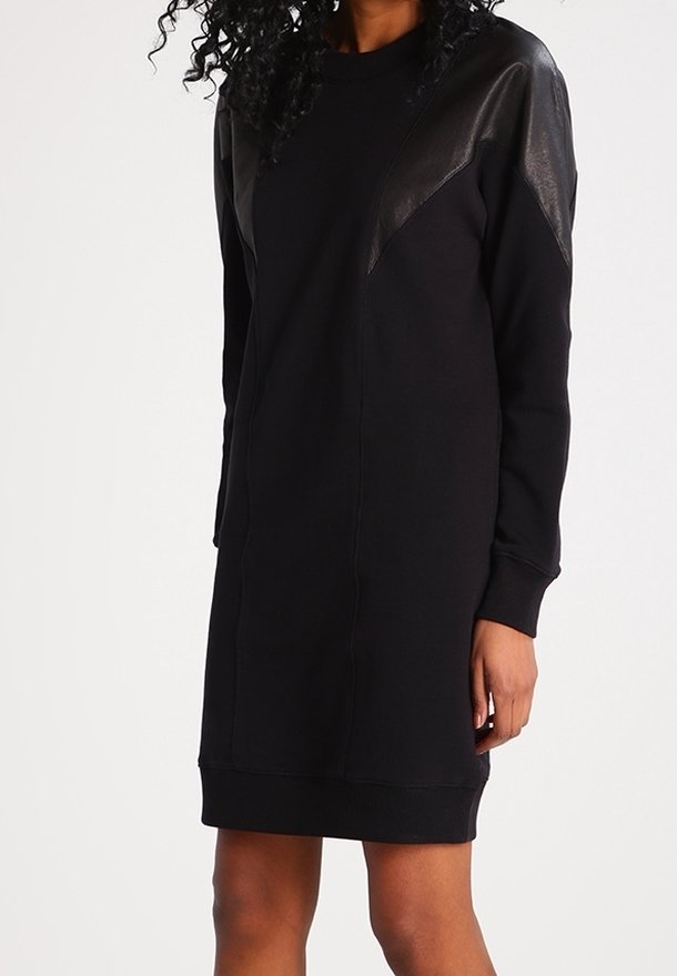 Femme portant une robe-pull noire mi-longue avec des épaules à panneaux en cuir et des manches longues, sur un fond clair uni.