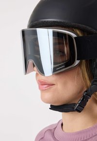 Gogle narciarskie z dużą, przyciemnianą szybą, czarnym paskiem i białą ramką. Noszone z czarnym kaskiem i fioletową, fakturowaną bluzką.