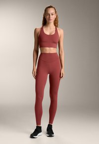 Červený atletický krátky top a legíny s vysokým pásom vyrobené z pružnej tkaniny, v kombinácii s čiernymi bežeckými topánkami, na neutrálom pozadí.