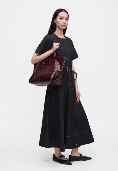 Femme en robe noire et chaussures noires à bouts pointus tenant un grand sac cabas en cuir rouge foncé brillant sur son épaule.