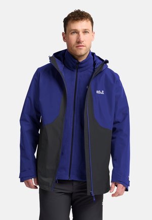 Man draagt een blauw-zwarte waterdichte jas met het Jack Wolfskin-logo, kijkt met een neutrale uitdrukking vooruit tegen een effen achtergrond.