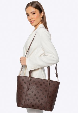 Žena v bílém saku drží velkou hnědou koženou kabelku s vzorem přes rameno na jednoduchém pozadí.