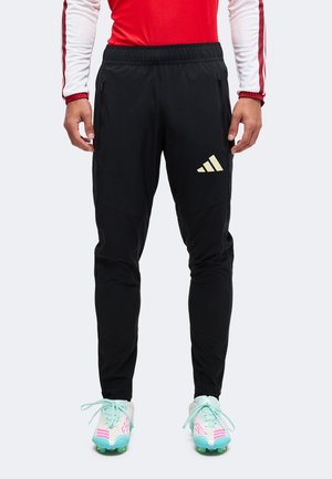 Άτομο που φοράει μαύρη φόρμα Adidas, κόκκινο-λευκό μπλουζάκι με μακριά μανίκια και λευκά παπούτσια ποδοσφαίρου με τιρκουάζ κορδόνια και ροζ λεπτομέρειες.