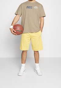 Camiseta del logo de la NBA de color naranja, pantalones cortos de baloncesto amarillos, zapatillas blancas, sosteniendo un balón de baloncesto Spalding rojo. Ropa atlética básica.