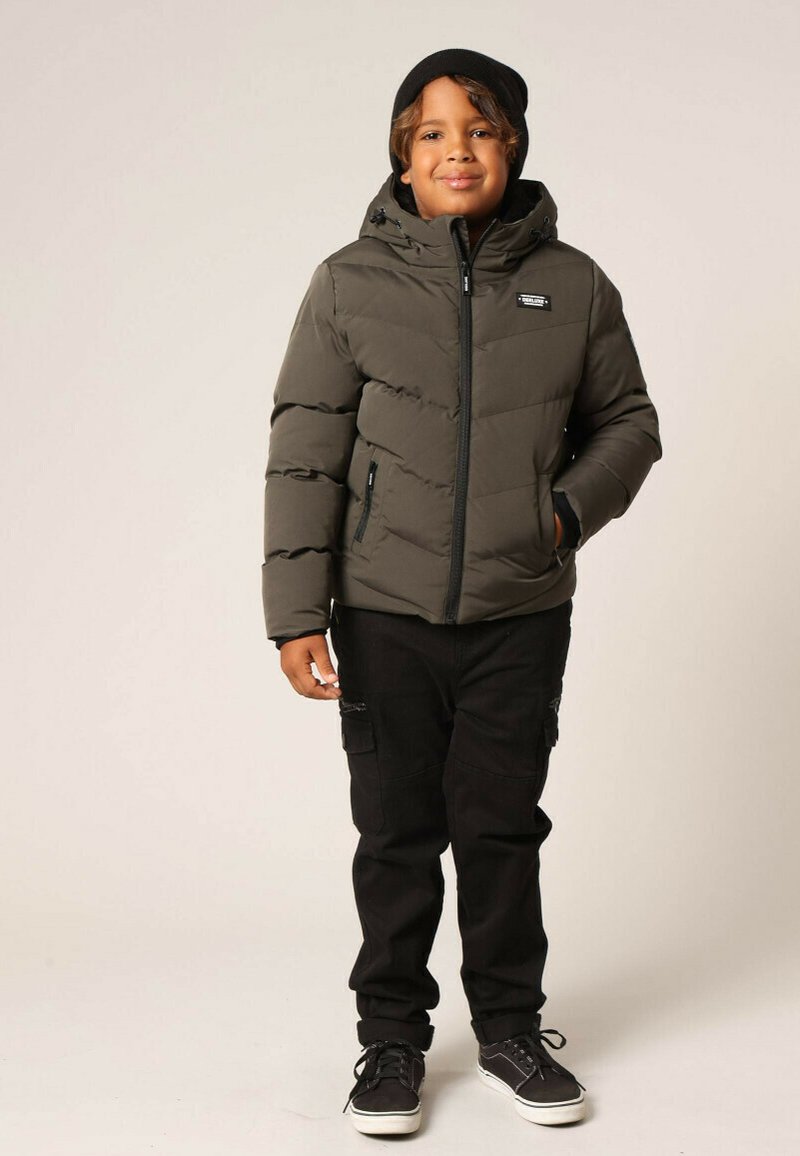 Veste rembourrée olive avec capuche, fermeture éclair à l'avant et poches sur les côtés ; portée avec un pantalon cargo noir et des baskets noires.