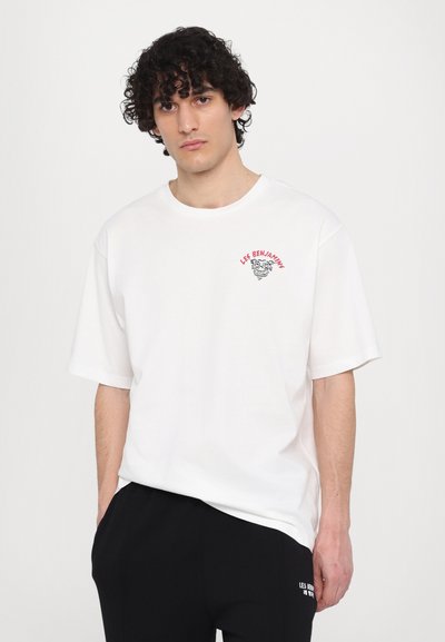 T-shirt en coton blanc avec un col rond et des manches courtes. Présente un imprimé graphique rouge sur la poitrine gauche indiquant "LES BENJAMINS."