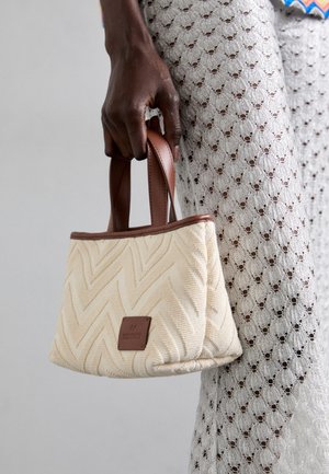 Cremfarbener gewebter Handtasche mit Chevron-Muster, braunen Ledergriffen und einem kleinen gebrandeten Lederanhänger. Texturierte Stoffoberfläche.