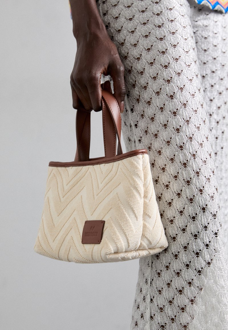 Sac à main tissé couleur crème avec un motif en chevrons, poignées en cuir marron et une petite étiquette en cuir de marque. Surface en tissu texturé.