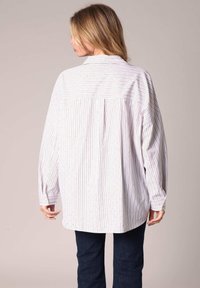 Woman with blonde hair wearing oversized white shirt with thin vertical stripes and dark jeans, standing facing away against neutral background.