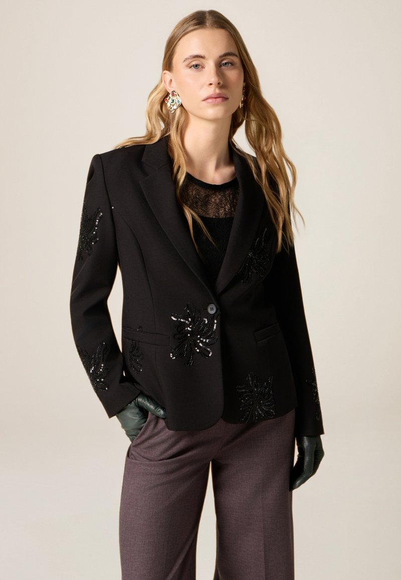 Zwarte blazer met kralenversieringen, twee zakken aan de voorkant en een enkele knoopsluiting. Draagt over een zwarte top van kant en gecombineerd met grijze broek.