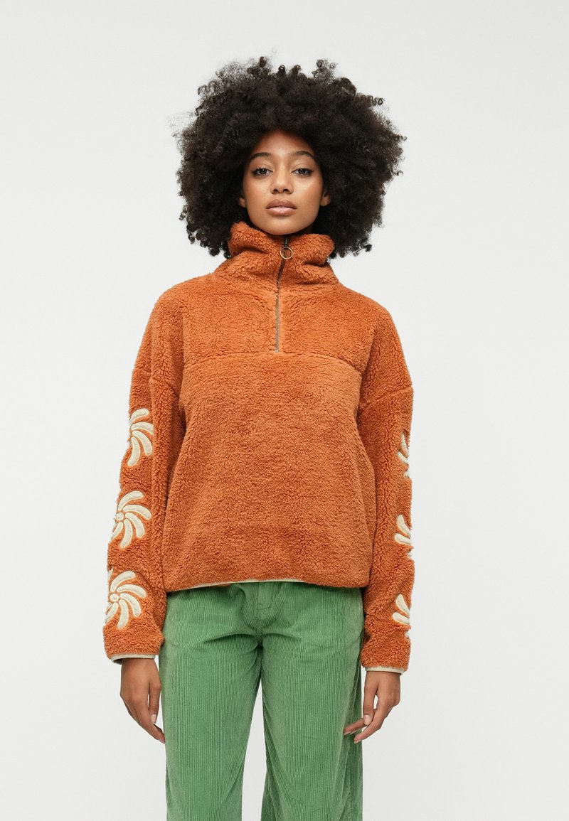 Hnedý chlpatský pulóver s vysokým golierom, polovičným zipsom a kvetinovou výšivkou na rukávoch. V kombinácii so zelenými kordovými nohavicami.