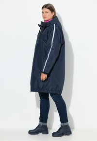 Mujer vestida con un impermeable con capucha de color azul marino con una franja reflectante en las mangas, jeans oscuros y botas negras hasta el tobillo, de pie de lado contra un fondo blanco.