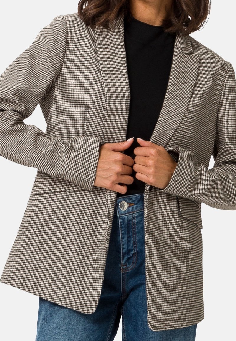 Personne portant un haut noir, un jean bleu et un blazer beige à motif pied-de-poule, tenant les revers du blazer avec les deux mains.