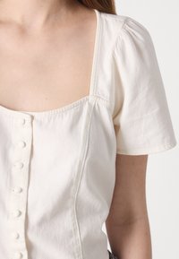 Blusa bianca a maniche corte con scollatura quadrata, chiusura frontale con bottoni e maniche leggermente a sbuffo, realizzata in un tessuto morbido.