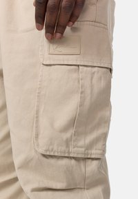 Beiga cargobyxor med en stor sidoficka, som har en texturerad tyg och en sydd logopatch nära midjan. Släta och hållbara material.