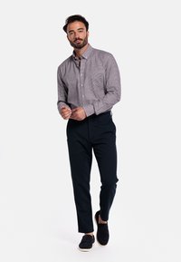 Man met een baard, gekleed in een grijs geruit overhemd, zwarte broek en zwarte instappers, staande tegen een witte achtergrond.