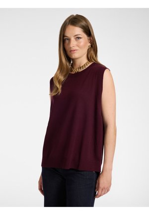 Mujer con cabello largo castaño claro que lleva una camiseta de punto sin mangas morada oscura, pantalones oscuros, collar de cadena dorada y pendientes de aro dorados.