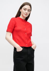 Czerwony krótki kardigan z krótkim rękawem, zapinany z przodu na guziki, z okrągłym dekoltem, wykonany z miękkiej dzianiny. Noszony na czarne jeansy z wysokim stanem.