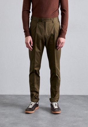 Olive green trousers with a tailored fit and pleated front, made of soft fabric. Paired with brown sneakers featuring white laces.