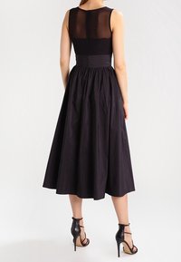 Abito nero senza maniche con schiena trasparente, vita aderente e gonna svasata. Tessuto liscio con zip nascosta. Abbinato a tacchi neri.