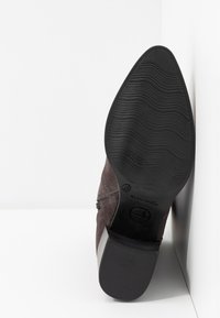 Chaussure en daim marron avec une semelle en caoutchouc noir. La semelle présente une texture rainurée et un bout arrondi. Accent élastique sur les côtés.