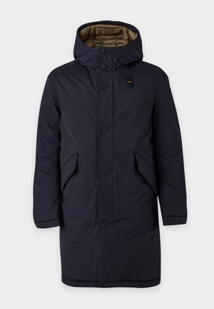 Un mannequin masculin se tient devant un fond uni et clair, vêtu d’un manteau bleu marine à capuche, d’un pull rayé, d’un pantalon beige et de bottes marron.