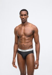 Μυώδης άνδρας στέκεται μπροστά σε απλό φόντο φορώντας μαύρα εσώρουχα Calvin Klein με λευκή λωρίδα στη μέση, στραμμένος προς τα εμπρός.