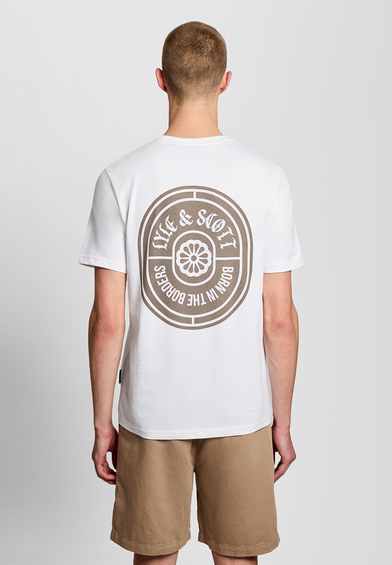 Άνδρας που φοράει λευκό T-shirt με κυκλικό μπεζ λογότυπο και κείμενο "Life & Soil Born in the Borders" στην πλάτη, συνδυασμένο με μπεζ σορτς.