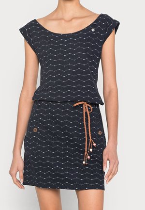 Kobieta ubrana w granatową sukienkę z białymi zygzakowatymi wzorami, krótkimi rękawkami, dwoma drewnianymi guzikami na spódnicy oraz oplecionym brązowym sznurem z koralikami jako paskiem.