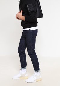 Tmavě modré džíny s úzkým střihem, doplněné černou mikinou s kapucí a kapsou na zip a bílými teniskami s šedými detaily.