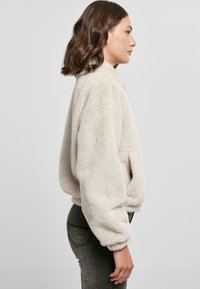 Fleece jakke i lyse beige med høj krave, sænkede skuldre, sidelommer og elastiske ærmer, parret med mørke grå jeans.