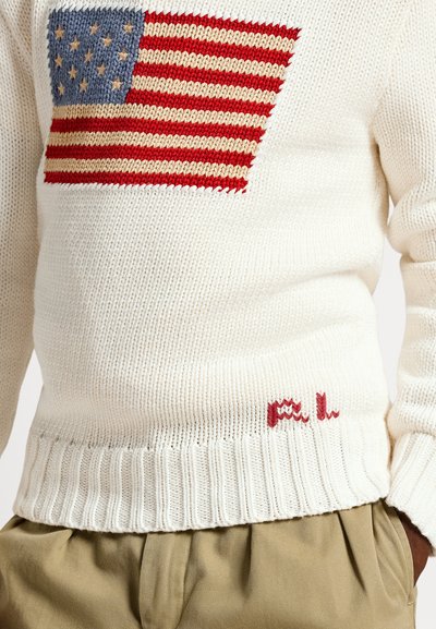Beli pleteni pulover z dizajnom ameriške zastave z rdečimi in modrimi poudarki, rebrastimi manšetami in izvezenimi inicialkami "P.L." na spodnjem robu.