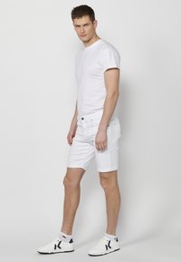 Camiseta blanca y pantalones cortos de mezclilla blanca con puños enrollados, combinados con zapatillas blancas acentuadas con detalles en azul marino. Fondo neutro.