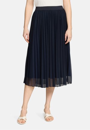 Kobieta ubrana w granatową plisowaną spódnicę midi z czarnym pasem w talii, białą bezrękawnikową bluzkę oraz białe sandały wsuwane.