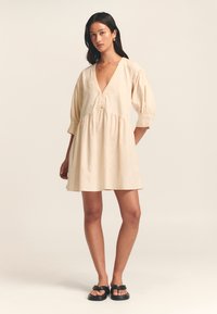Korte beige jurk met een diepe V-hals, pofmouwen en een geraffineerde taille, gemaakt van zachte stof. Bevat knoopaccenten bij de halslijn.