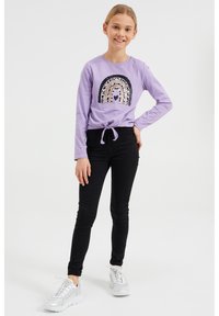 Fialová dlouhá halenka s duhovým flitrovým vzorem, kombinovaná s černými džíny a stříbrnými teniskami. Halenka má detail v podobě uzlu vpředu.