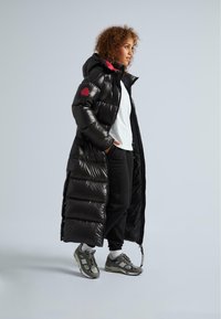 Giacca a vento nera con materiale lucido, lunghezza lunga, cappuccio e accenti rossi. Design a strati con trapuntatura prominente e chiusura frontale con zip.