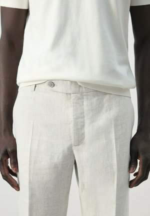 Hombre con pantalones texturizados de color beige claro con cierre de botones y camisa blanca de manga corta, de pie con las manos relajadas a los lados.