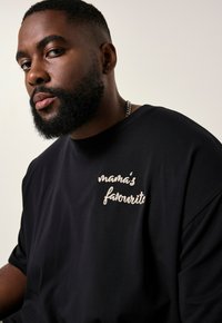 Sort bomulds t-shirt med korte ærmer, der har lyserøde, broderede bogstaver "mamas yndling" på venstre brystområde.