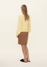 Żółty sweter dziergany z koronkowym wzorem i bufkami na rękawach, zestawiony z brązową prążkowaną spódnicą do kolan oraz czarnymi, puszystymi butami wsuwanymi.