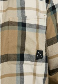 Tejido a cuadro en beige, negro y azul con un bolsillo cuadrado cosido y un pequeño parche negro que presenta un diseño minimalista en blanco.
