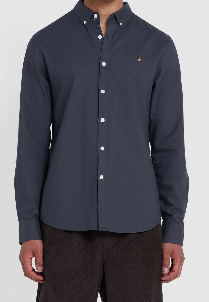 Homme portant une chemise boutonnée bleu marine foncé avec un petit logo brodé sur la poitrine et un pantalon marron, debout sur fond blanc.