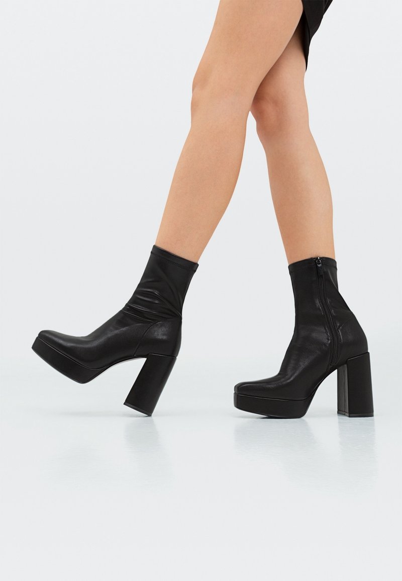 Jambes portant des bottines à plateforme en cuir noir avec talons blocs et fermetures éclair latérales, marchant sur un sol clair et réfléchissant.