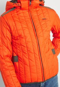 Světlý oranžový prošívaný kabát s šedým zipem, kapucí a akcenty kapes, nošený přes modré džíny.