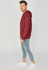 Bordowa bluza z kapturem z sznurkiem, z naszywką logo na rękawie. Połączona z podartymi jasnoniebieskimi dżinsami i białymi trampkami.