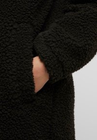 Cappotto in pile nero con materiale testurizzato e morbido; presenta una tasca e ampidi polsini morbidi; primo piano che mostra il dettaglio del tessuto e la posizione della mano.
