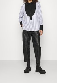 Gestreifte Bluse in Weiß und Blau mit einem schwarzen Kragen und Brustbereich, kombiniert mit schwarzen Lederhosen und klobigen schwarzen Stiefeln.