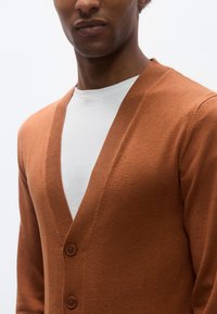 Καφέ πλεκτή ζακέτα με V-λαιμόκοψη, διαθέτει τρία κουμπιά και λεία υφή, φορεμένη πάνω από λευκό μπλουζάκι με στρογγυλή λαιμόκοψη.