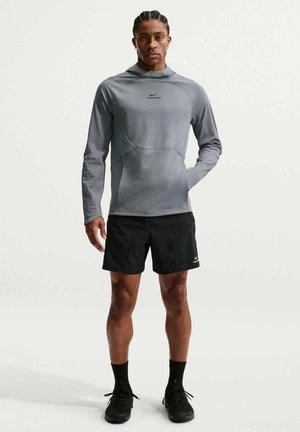 Mann steht, trägt einen grauen Nike Pro Hoodie mit langen Ärmeln und schwarze Nike Pro Sportshorts mit schwarzen Schuhen und Socken vor einem einfarbigen Hintergrund.