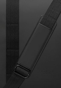 Zaino nero in materiale liscio, con spallacci in tessuto regolabili e una toppa rettangolare con logo, che enfatizza un design minimalista.
